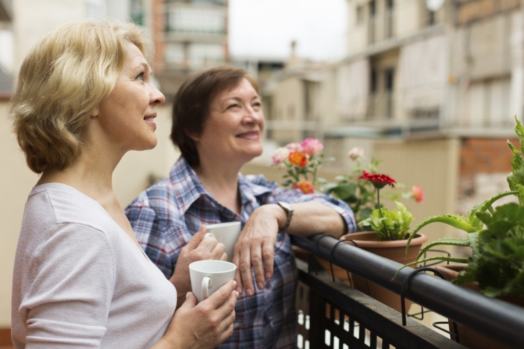 Smiling aged female friends relaxing with cup of coffee on balcony